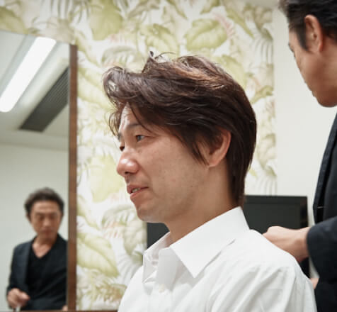 前髪が長めの男性が黒いシャツの男性にヘアスタイルを整えてもらっている